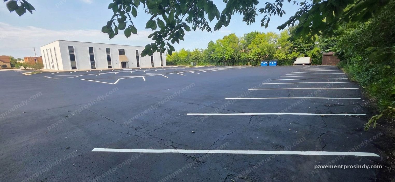 A single-storey office building's parking lot striped in white — angled stalls across the near half and straight rows along the tree line, over a sealed surface that still shows cracking, with two blue dumpsters at the far corner.