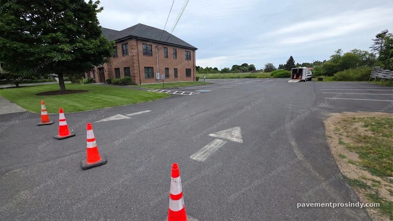 A commercial parking lot before restriping — the drive-lane arrows worn down to faint grey outlines, stall lines faded across the lot, and safety cones staged in the foreground under an overcast sky.