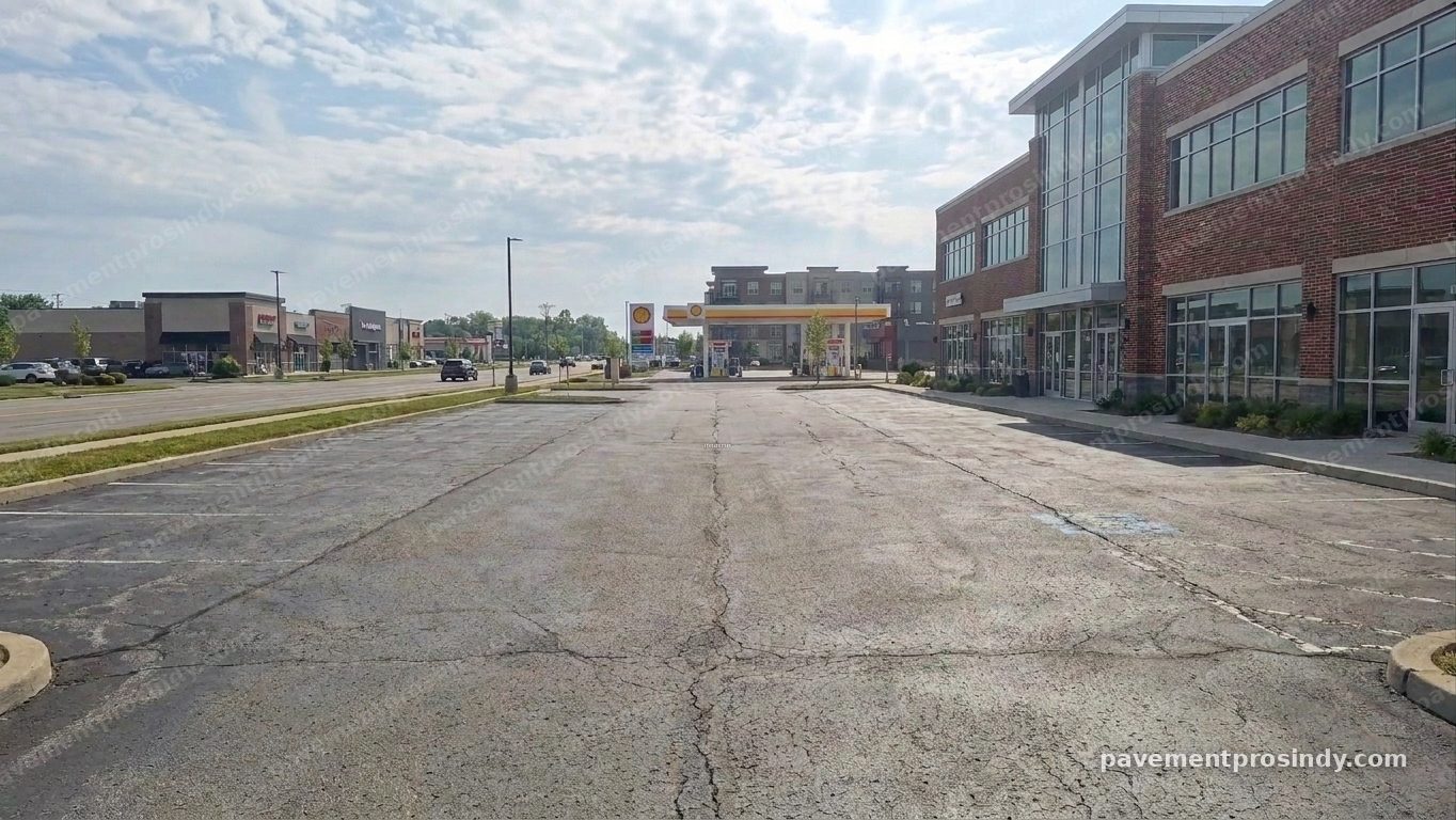 A retail center parking lot before work — the asphalt faded to pale grey, cracks running the length of the surface and across the stalls, and stall lines worn down to faint marks, with a gas station and storefronts across the street.