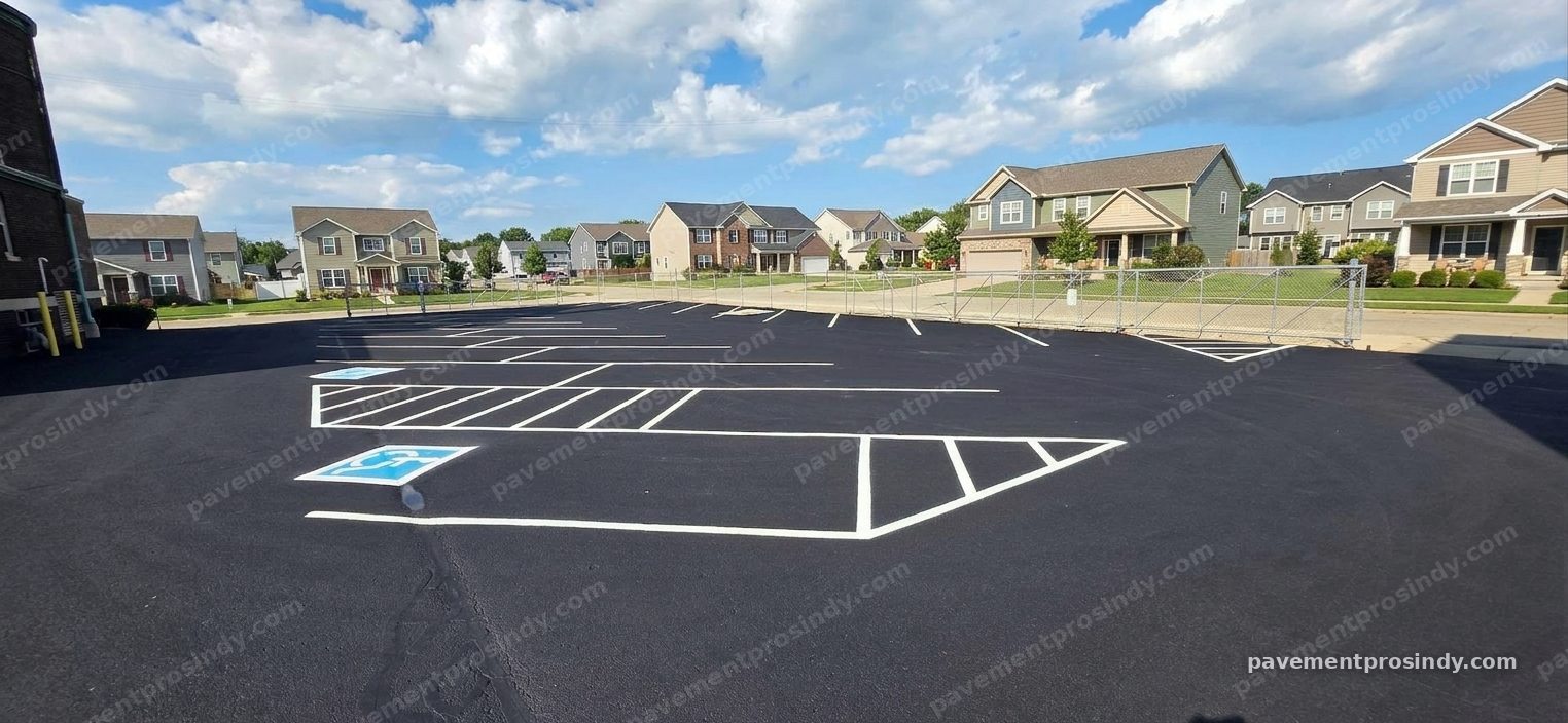 A freshly sealed parking lot striped in white — two accessible spaces marked with blue wheelchair symbols and hatched access aisles alongside, rows of stalls across the lot, and a striped triangular island at the drive-lane end, with houses across the street beyond the fence.